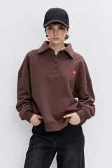 Sudadera de algodón color marrón, con cuello polo y cierre frontal de botones. Presenta un diseño holgado, mangas largas con puños elásticos y un pequeño parche bordado en forma de corazón en el pecho.