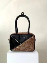Cartera rígida negra con detalles en rojo y tachas plateadas. Tiene herrajes y cierre metálico plateado marca YKK, asa fina para llevar al hombro o en la mano y correa larga extra regulable. El interior es forrado, tiene un bolsillo con cierre enmarcado en cuero, y un segundo bolsillo plaqué.