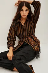 Saco tipo camisa de algodón con rayas verticales marrones y negras. Presenta bordados de cuentas en tonos tierra en el hombro izquierdo y se ajusta a la cintura con un lazo de viscosa estampado en tonos naranjas y negros.