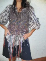 Top de diseño híbrido que combina un cuerpo de tejido de punto con brillo multicolor y mangas de camisa a cuadros. Presenta un ruedo con flecos largos decorativos y cordones frontales.