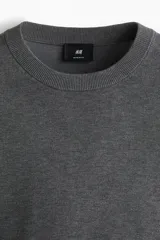 Buzo de punto fino color gris oscuro, de manga larga y cuello redondo, con remates acanalados en cuello, puños y bajo.