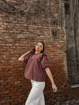 Blusa color chocolate con mangas cortas abullonadas, cuello redondo con volados y detalles de costura en zigzag de color rojo en el pecho y volados oscuros en los hombros.