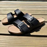 Sandalia tipo Birkenstock con doble tira ajustable de cuero sintético negro y hebillas metálicas. Cuenta con plantilla anatómica de corcho y suela de goma.