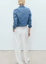 Chaqueta de jean azul de corte crop, con cuello camisero, cierre frontal de botones metálicos, bolsillos con solapa en el pecho y mangas largas con puños abotonados.