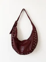 Cartera estilo hobo de cuero sintético color bordó, con diseño de media luna y detalles de ojales metálicos plateados a lo largo de los bordes. Cuenta con correa ajustable y cierre superior.