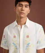 Camisa blanca de lino y algodón, de corte comfort, cuello inglés y abotonado frontal. Presenta manga corta y un bordado floral multicolor con motivos de aves y flores.