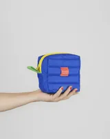 Neceser de viaje acolchado color azul, con cierre superior amarillo y tirador lateral verde. Presenta una etiqueta rectangular naranja con el logo de la marca en el frente.