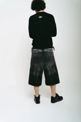 Bermuda de jean color negro con lavado acid wash gris. Presenta un corte baggy y detalles bordados en plateado y blanco que simulan ondas o llamas en la parte frontal superior e inferior.