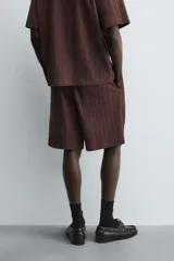 Bermuda de corte relajado color marrón oscuro, confeccionada en tejido jacquard con textura acanalada vertical. Presenta cintura elástica ajustable con cordón, bolsillos delanteros y un bolsillo trasero de plastrón.