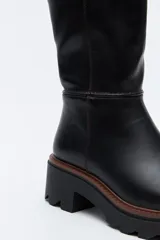 Bota de caña media en cuero sintético color negro, con diseño minimalista y suela robusta dentada con detalle de borde en color marrón.