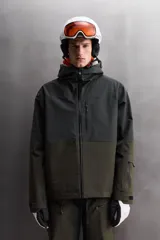Cazadora deportiva de esquí con diseño bicolor en tonos negro y verde oliva. Presenta capucha amplia con visera, cierre frontal con cremallera termosellada, bolsillos frontales y en manga con cierres impermeables, y aberturas de ventilación en las axilas. Incluye faldón interior antinieve, puños ajustables y tecnología de rescate RECCO.