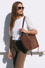 Bolso tipo shopper de gamuza vegana color marrón, con tiras colgantes decorativas a los lados y placa metálica frontal con el logo de la marca. Cuenta con asas largas de hombro.