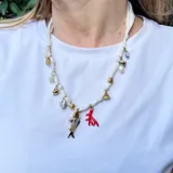 Collar corto hecho con cinta de tela blanca y dijes dorados, rojos y nacarados con forma de pez, corazón, coral, flor y cuentas.