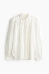Blusa oversize blanca de algodón y viscosa, con cuello camisero, tapeta de botones ocultos y detalles fruncidos en el canesú delantero y trasero. Presenta mangas globo largas con puños abotonados.