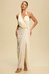 Vestido largo de fiesta color crema, con diseño drapeado, escote halter y espalda descubierta. Presenta un detalle metálico dorado en el hombro y una abertura frontal en la falda.