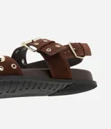 Sandalia plana tipo sport con tiras de efecto gamuza en color marrón. Presenta cierre con hebillas metálicas y detalle de tachuelas y ojetes dorados en las tiras.