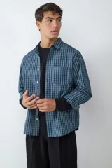 Camisa de corte oversize con estampado de cuadros en tonos azules y verdes, confeccionada en algodón. Presenta cuello clásico, cierre frontal con botones, mangas largas con puños abotonados y un bolsillo aplicado en el pecho.