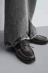 Zapato náutico de cordones, color negro, fabricado en piel con pespuntes a contraste en color claro. Presenta una horma redonda y suela track gruesa.