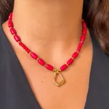 Collar compuesto por cuentas rojas de diferentes formas, incluyendo cuentas cilíndricas lisas y pequeñas cuentas facetadas, intercaladas con separadores dorados. Presenta un dije central dorado con una forma orgánica e irregular.