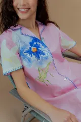 Vestido corto de lino y algodón color rosado, con cuello de solapa y manga corta. Presenta un diseño abotonado al frente con estampados de flores azules y saltamontes verdes, además de detalles de bordado a mano en contraste azul en los bordes.
