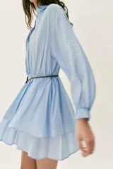 Vestido corto de color celeste con cuello solapa, cierre frontal de botones y manga larga con puños elásticos. Presenta cintura elástica, falda con volantes y un cinturón fino con un pequeño bolsillo de flecos incorporado.