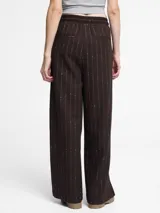 Pantalón de vestir de corte ancho y tiro alto, color marrón oscuro, con rayas diplomáticas finas en hilo metálico. Presenta pliegues frontales y cintura con elástico y cordón ajustable.