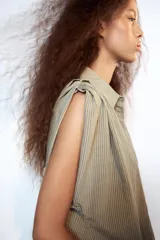 Camisa fluida de cuello solapa y manga corta, con diseño a rayas en tonos beige y blanco, detalle de pliegues en los hombros y cierre frontal con botones.