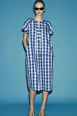 Vestido midi de Zara Woman Collection confeccionado en mezcla de viscosa, con estampado de cuadros en tonos azul y celeste. Presenta cuello redondo, manga corta, bolsillos ocultos en las costuras y cierre frontal con cordones entrelazados.