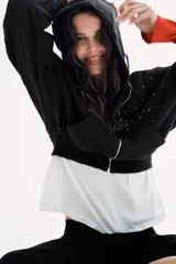 Sudadera negra con capucha, cierre frontal de cremallera metálica y mangas largas con puños. Presenta un diseño corto con bolsillo tipo canguro y detalle de texto 'California' aplicado con brillos en el frente.