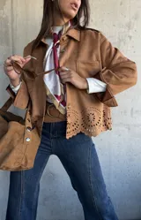 Chaqueta de gamuza color camel con cierre frontal de botones, cuello clásico y detalle de calados decorativos en el ruedo. Presenta bolsillos frontales y mangas largas con puños.