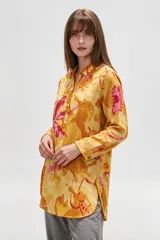 Camisa de lino larga y holgada, de color amarillo con estampado abstracto en tonos fucsia y naranja, con cuello camisero, cierre frontal parcial con botones y aberturas laterales en el dobladillo.