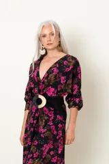Vestido midi de seda natural con estampado floral en tonos fucsia sobre base negra. Presenta un diseño drapeado con escote en V cruzado, mangas largas abullonadas y una abertura frontal en la falda.