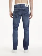 Jeans de hombre de mezclilla azul oscuro con efecto lavado y arrugas marcadas en la parte delantera. Presentan un corte slim, tiro alto, cierre de cremallera y botón, y diseño clásico de cinco bolsillos. Incluyen un parche con logo en la cintura trasera.