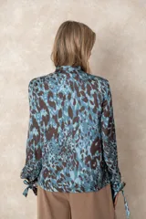 Camisa de seda con estampado animal print en tonos azules y marrones. Presenta cuello con lazo, cierre frontal con botones forrados y mangas largas con puños ajustables mediante lazos.