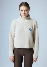 Sweater de punto grueso color crema, con cuello alto y parche circular con bordado en el pecho.