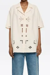 Camisa de manga corta color crema, con bordado inglés en el delantero y las mangas, cuello con solapas y cierre frontal con botones.