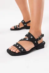 Sandalia baja de cuero negro con tiras cruzadas en la capellada decoradas con tachas metálicas plateadas. Cuenta con cierre de hebilla en el talón, suela plana con pequeño tacón y punta cuadrada.