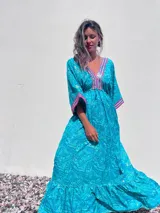 Túnica larga de estilo boho, color turquesa con estampado paisley en tonos azules y dorados. Presenta escote en V profundo con bordados decorativos en color fucsia y blanco, mangas amplias tipo kimono y ajuste en la cintura con cordón. El diseño incluye un ruedo con volado y terminaciones con borlas.