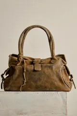 Cartera de ante color marrón con terminación gastada, cuerpo blando, doble asa de mano, cierre frontal con hebilla y detalles de tachas. Incluye cordones de ajuste en los laterales y un colgante metálico decorativo.