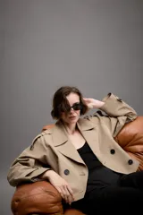 Trench corto de color beige con cuello de solapa, cierre frontal cruzado con botones y mangas largas con trabillas ajustables en los puños. Presenta un diseño estructurado con hombreras y bolsillos delanteros de vivo.