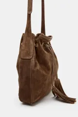 Bolso tipo bandolera de formato mini, confeccionado en piel de ante color marrón. Presenta un diseño de saco con cierre mediante cordón fruncido y detalle de borlas. Incluye una correa larga ajustable y herrajes metálicos dorados.