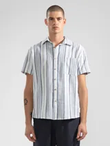 Camisa de manga corta a rayas verticales azules y blancas, con cuello clásico, corte cuadrado y calce relajado. Presenta botones de coco con logo y parche distintivo de Polonio. Confeccionada en Italia con mezcla de lino y algodón.
