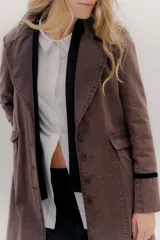 Tapado largo de jean azul oscuro, con cuello solapa y puños con detalle de cinta de terciopelo negro. Presenta costuras visibles que definen paneles y un logo bordado en la espalda.