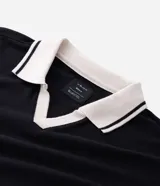 Remera de corte boxy en tejido crepé, color negro, con cuello polo en contraste color blanco y mangas cortas. Presenta un diseño holgado y moderno con textura sutil en la superficie.