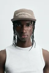 Gorra de seis paneles color marrón topo con visera plana y ojales bordados. Presenta bordado frontal en blanco con el texto "Alternative Lifestyle" y "Trastevere, Rome.".