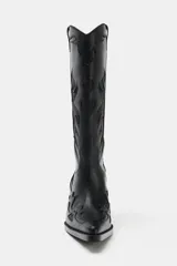 Bota estilo cowboy de caña alta, color negro, con detalles en relieve y costuras decorativas. Presenta punta fina, tiradores laterales y taco cuadrado inclinado.