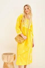 Vestido largo hasta los tobillos de color amarillo, con bordados florales blancos a contratono. Presenta mangas tres cuartos, escote redondo con cartera de botones frontales y un diseño holgado.