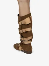 Bota de montar de caña alta y diseño holgado, confeccionada en cuero con acabado de gamuza color marrón. Presenta múltiples correas de cuero liso en color beige con hebillas metálicas doradas a lo largo de la caña y el empeine. Posee un taco bajo de aproximadamente 2,5 cm.