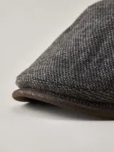 Boina estilo irlandés en color gris con textura jaspeada. Presenta un detalle lateral en cuero marrón con un botón metálico decorativo y visera integrada.