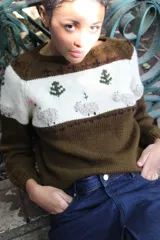 Sweater tejido a mano en punto jersey, color marrón con franja superior en color natural y cuello redondo. Presenta bordados artesanales en relieve de ovejas, árboles y flores en el pecho.